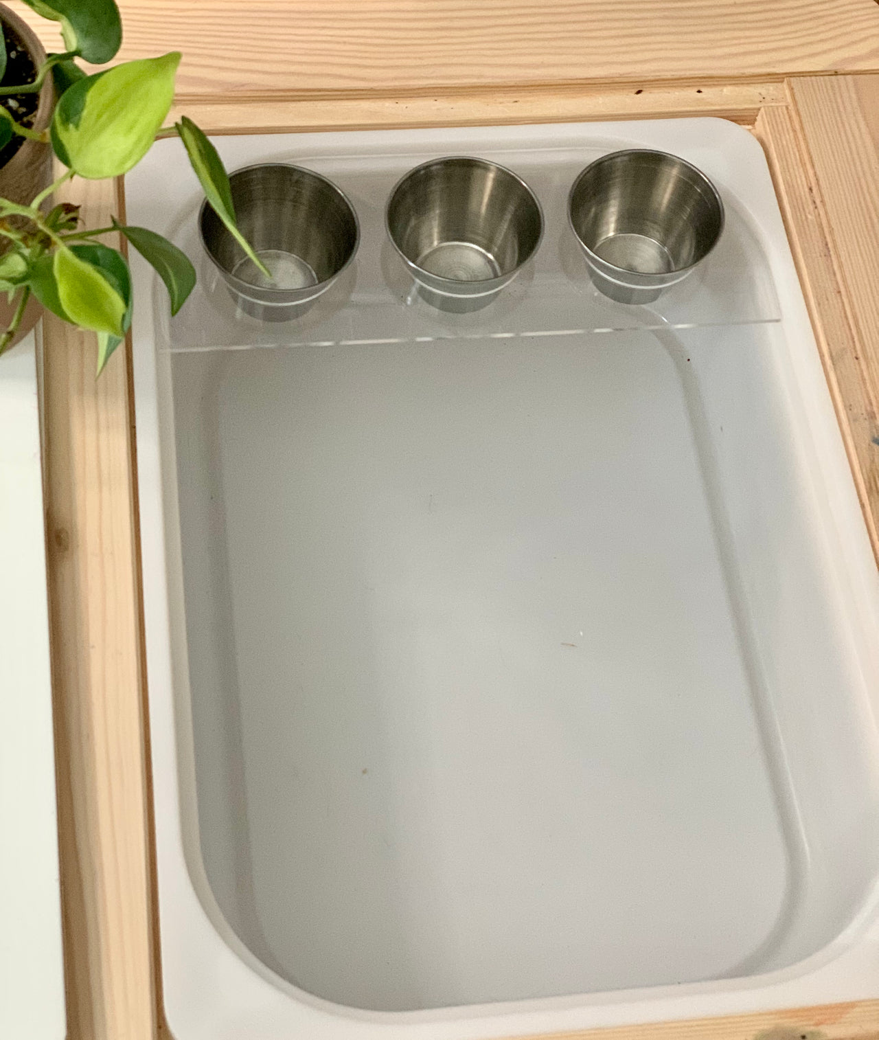 Edge piece Flisat table top insert with three stainless steel cups set in light wooden frame with green plant nearby