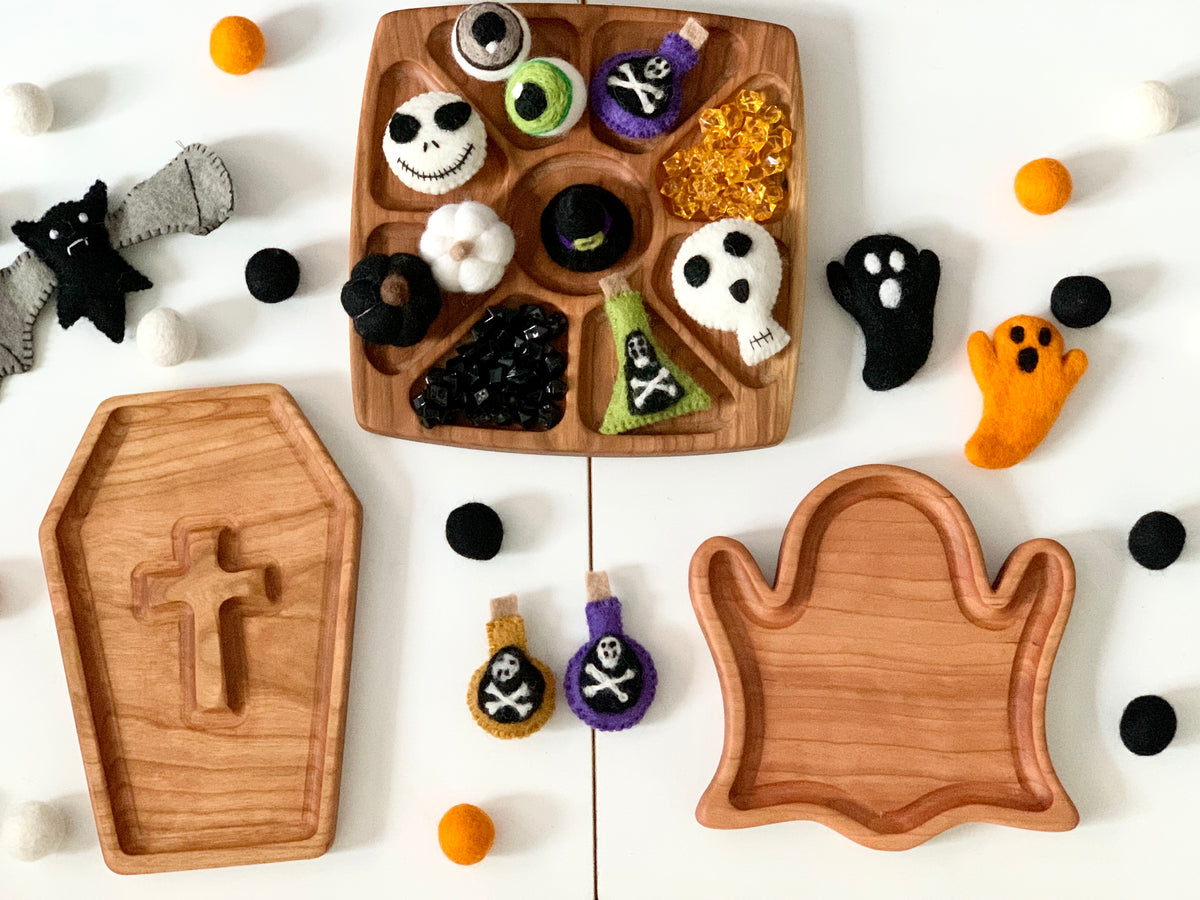 Wooden ghost and coffin-shaped sensory trays with felt Halloween decorations and black, orange, and white felt balls on white surface