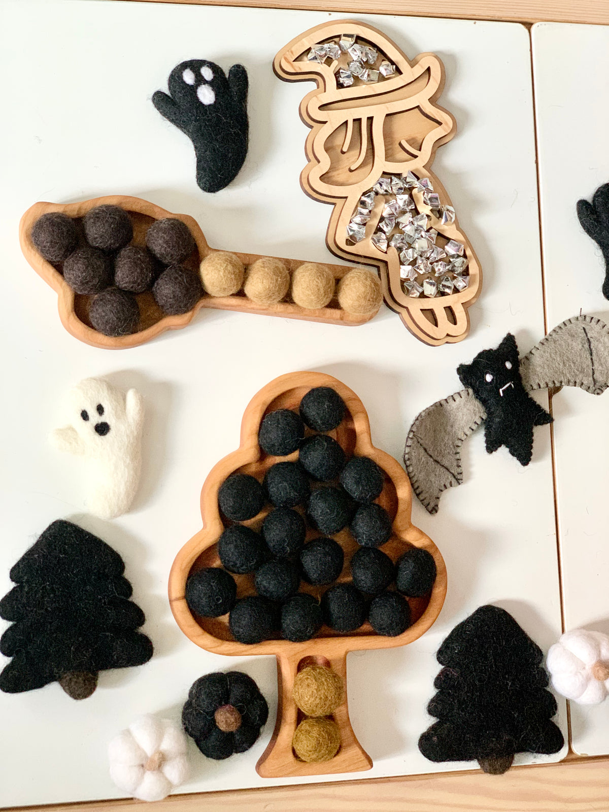 Wooden witch broom and paint brush sensory trays with felt balls, felt ghosts, bats, pumpkins, and trees on white table