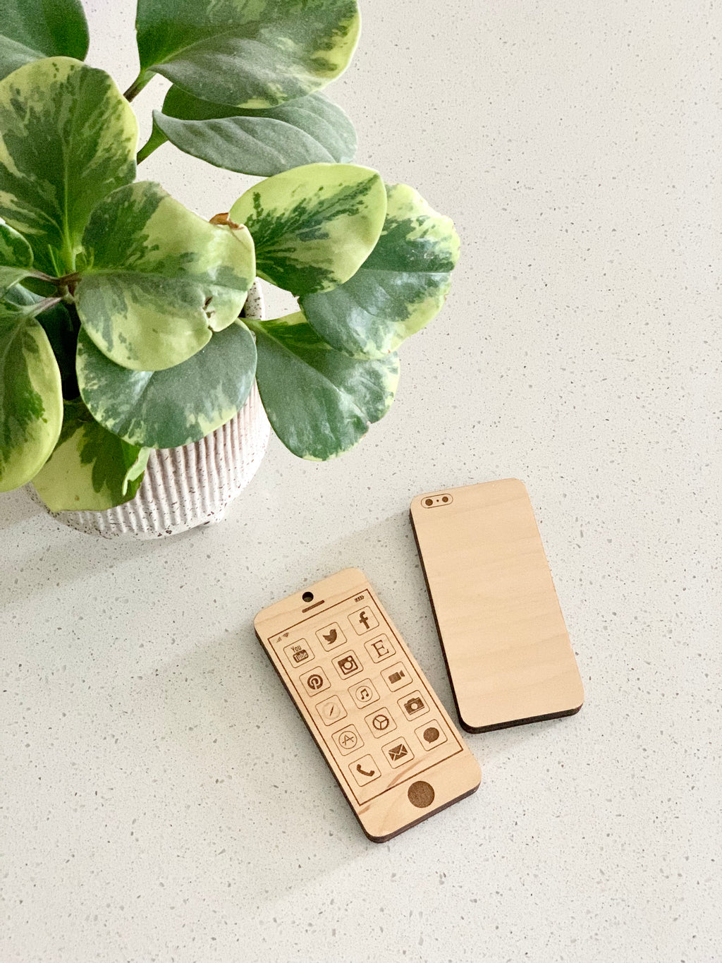 Two laser cut wooden toy phones with app icons and camera details on beige speckled surface next to green potted plant