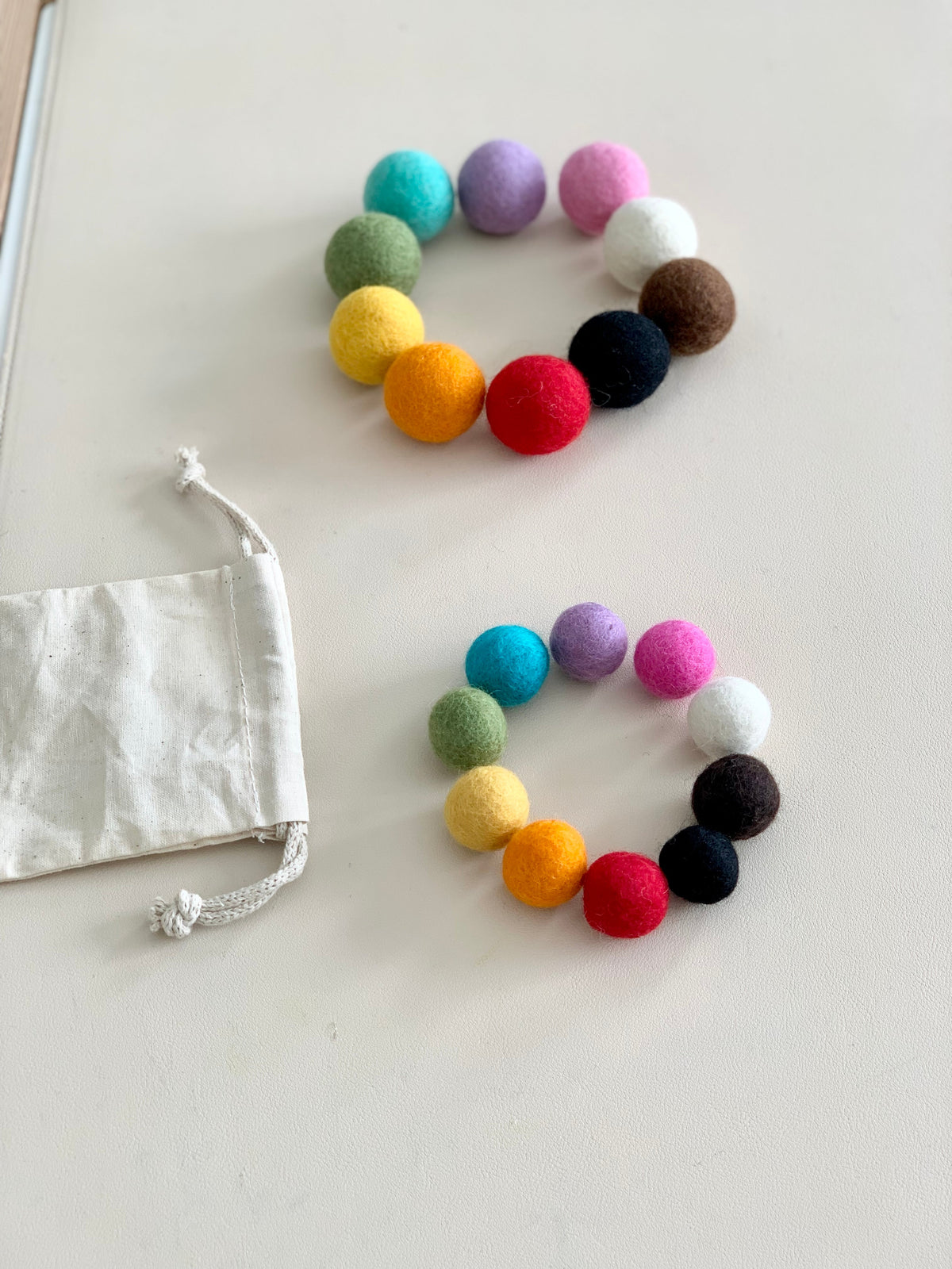 Two circles of colorful felt balls in large and small sizes with a natural cotton drawstring bag on white surface