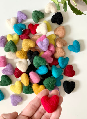 Small handmade felt hearts in multiple colors scattered on white surface with a hand holding one red heart
