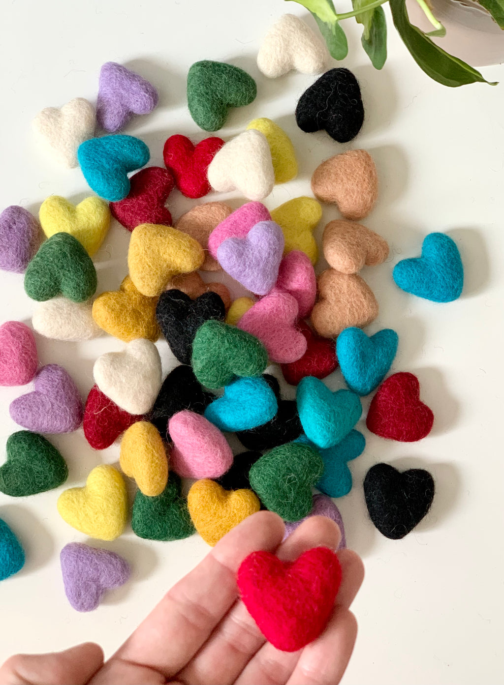 Small handmade felt hearts in multiple colors scattered on white surface with a hand holding one red heart