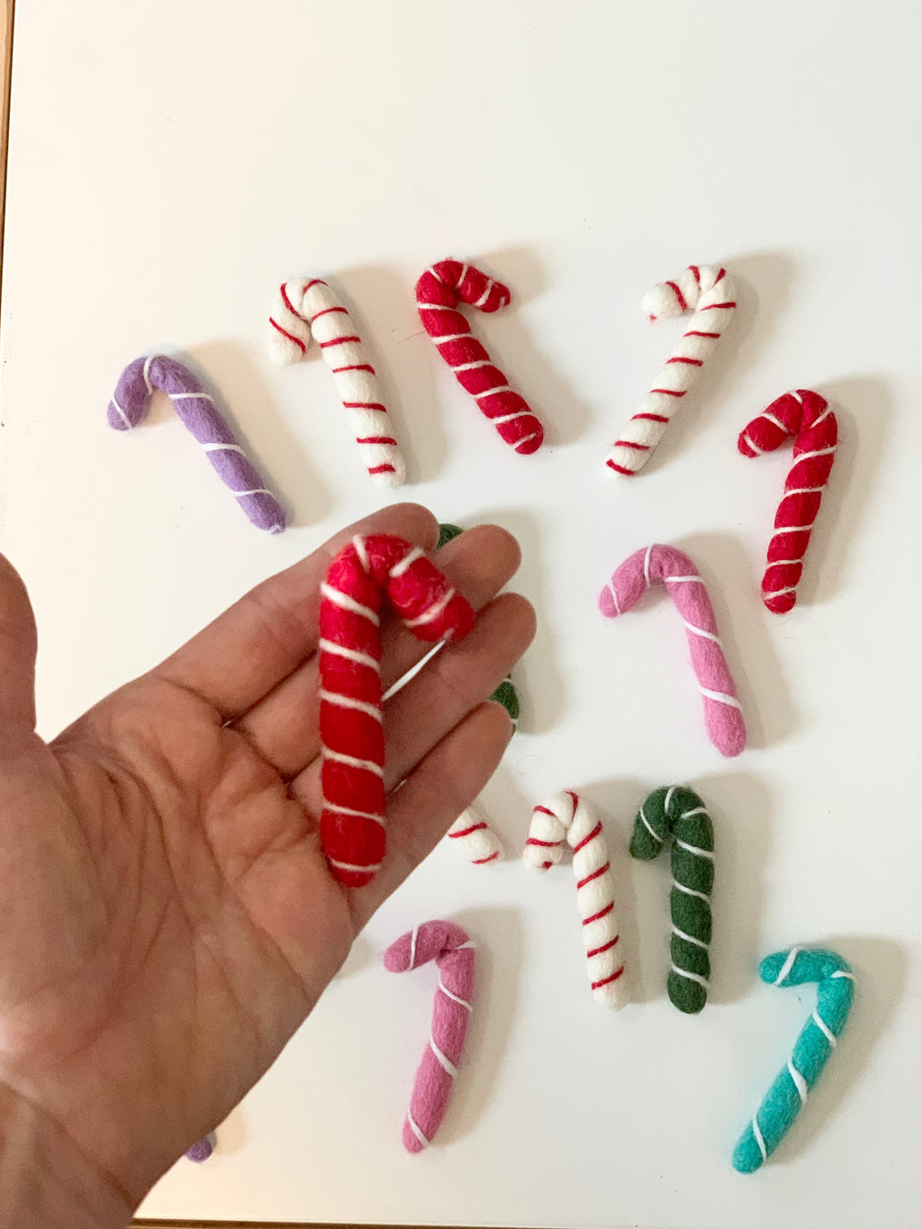 Hand holding a single red and white striped felt candy cane surrounded by assorted colored felt candy canes on white surface