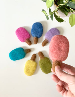 Handmade colorful felt popsicles in various shades arranged in a circle with one held in hand on white table
