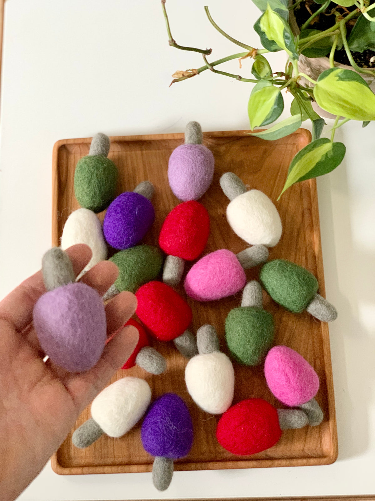Hand holding single purple felt light bulb with multiple colorful felt light bulbs on wooden tray near green plant on white table