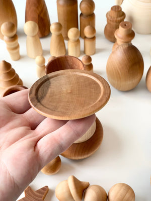 Hand holding small round wooden dish with various unfinished wooden peg dolls and shapes on a white surface
