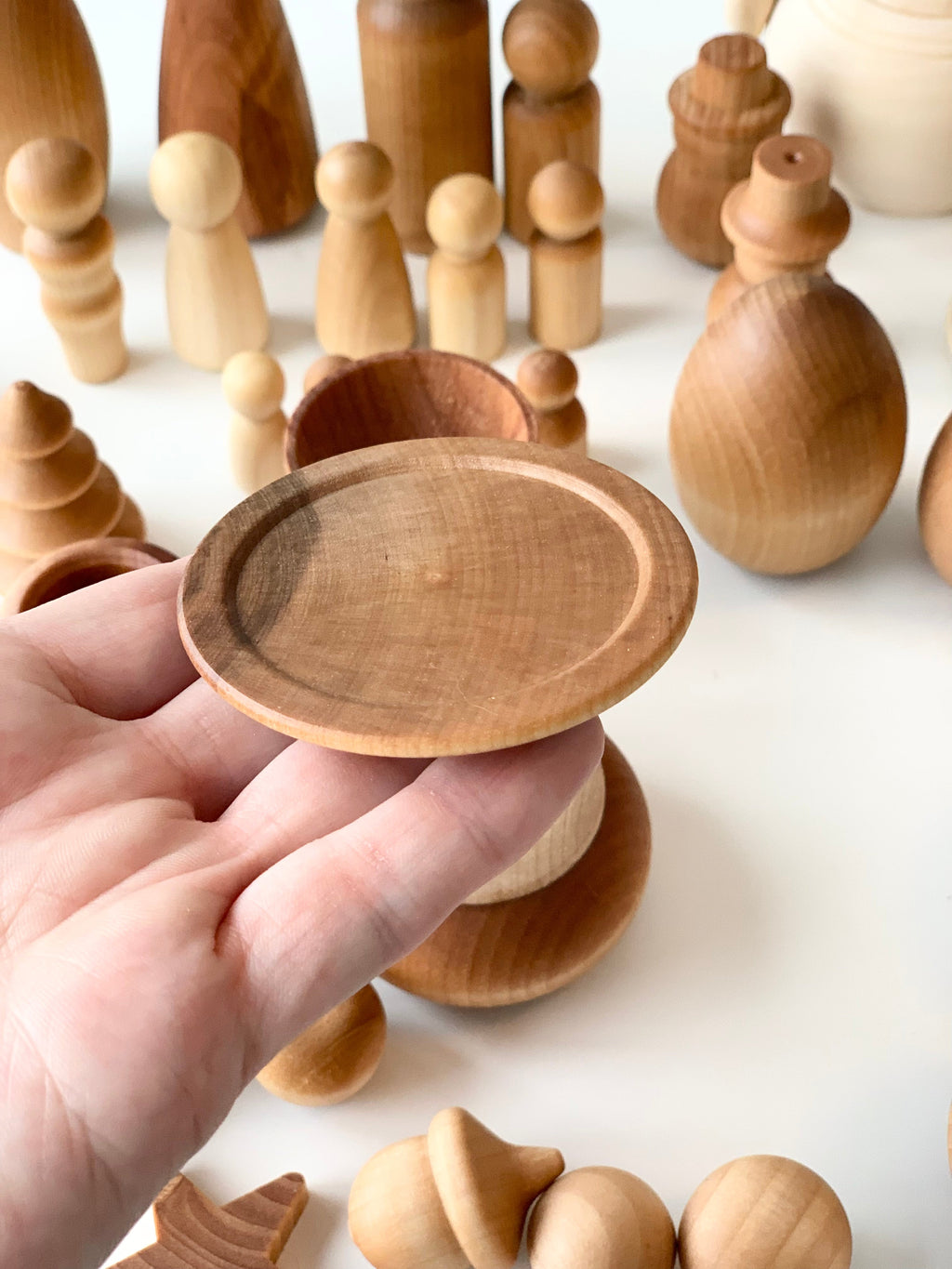Hand holding small round wooden dish with various unfinished wooden peg dolls and shapes on a white surface