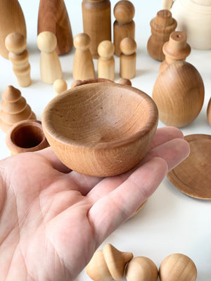 Hand holding shallow, round wooden bowl with various unfinished wooden figurines and shapes in the background