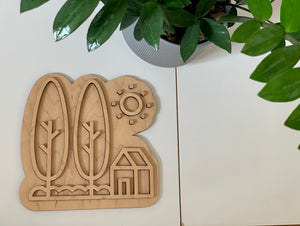 Maple plywood fillable house play tray with trees, sun, and house outline on white surface next to green plant
