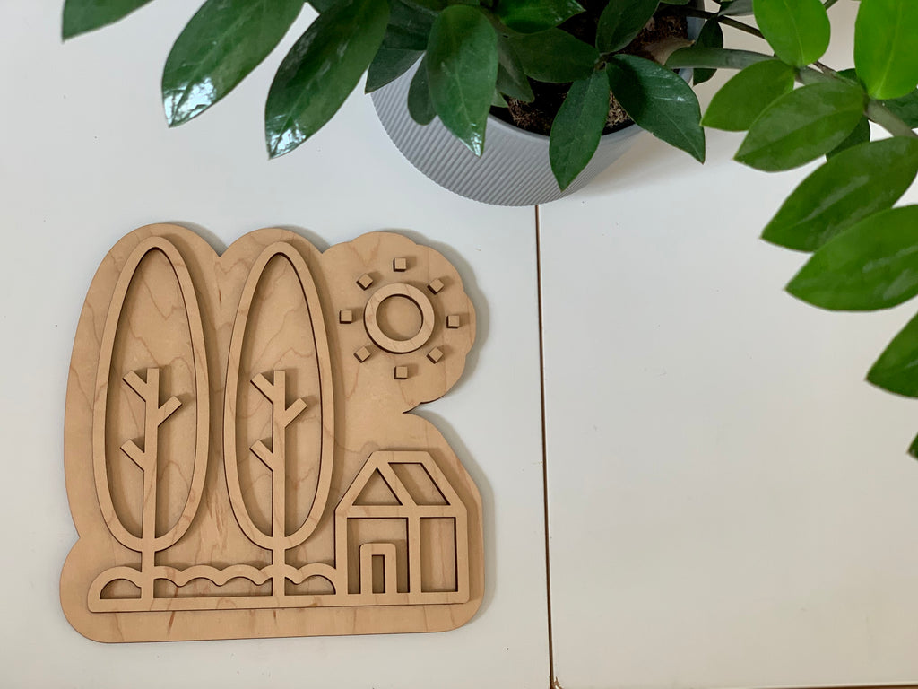 Maple plywood fillable house play tray with trees, sun, and house outline on white surface next to green plant