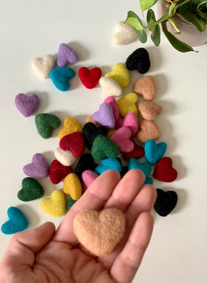 Assorted small handmade felt hearts in various colors scattered on white surface with hand holding peach heart