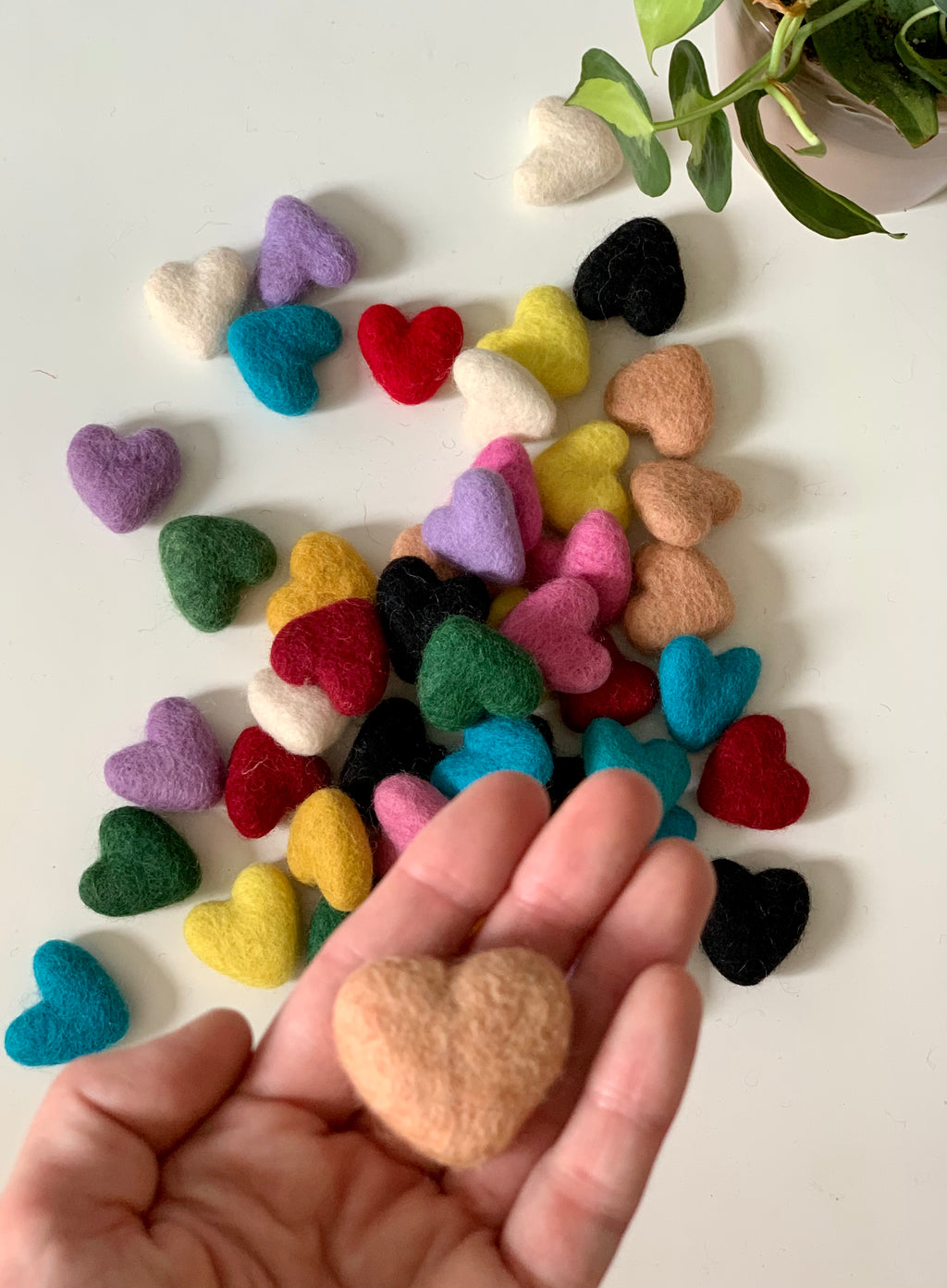 Assorted small handmade felt hearts in various colors scattered on white surface with hand holding peach heart