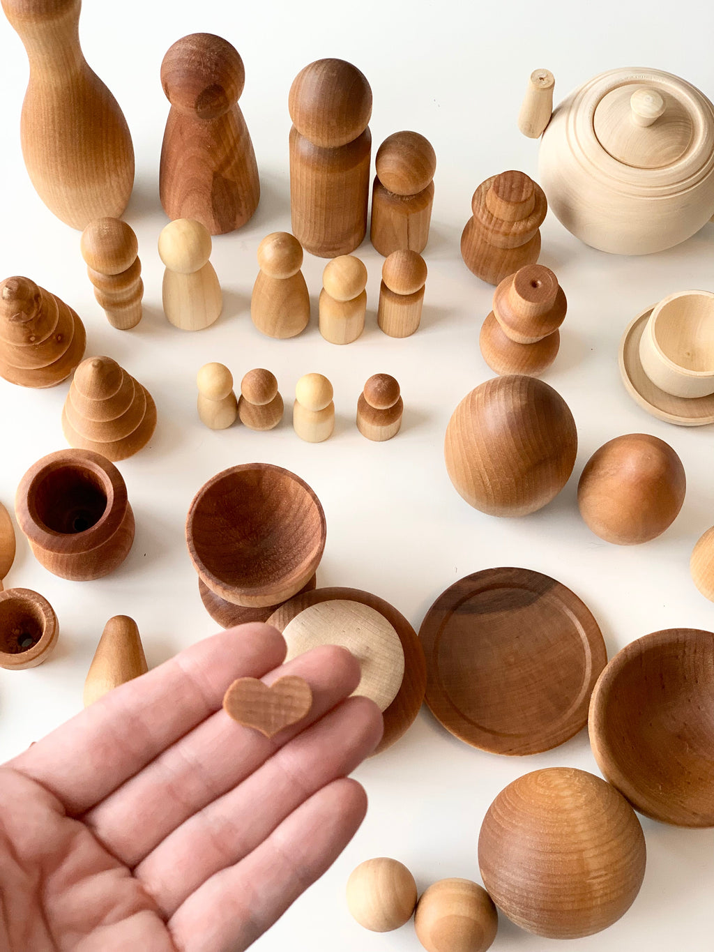 Hand holding a small natural wood heart shape with various smooth wooden figures, vessels, and toys on white surface