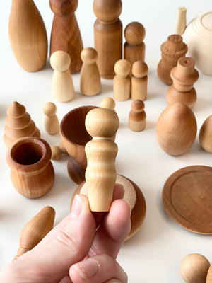 Hand holding small smooth natural wood peg doll with assorted handcrafted wooden bowls and shapes on white surface
