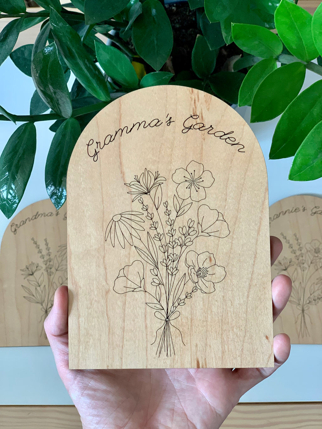Hand holding wooden personalized garden plaque engraved with wildflowers and "Gramma’s Garden" text with green leaves background