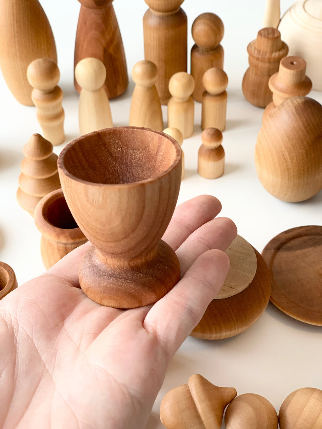 Hand holding smooth, unfinished wooden cup with various natural wood peg dolls and shapes on a white background