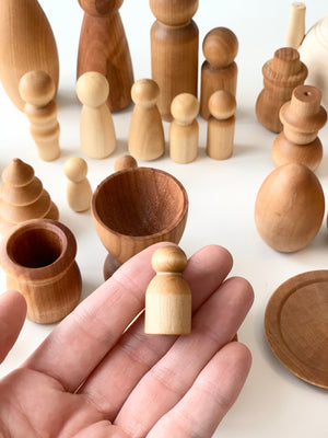 Hand holding small unfinished wooden peg doll among various natural wood shapes and vessels on white surface