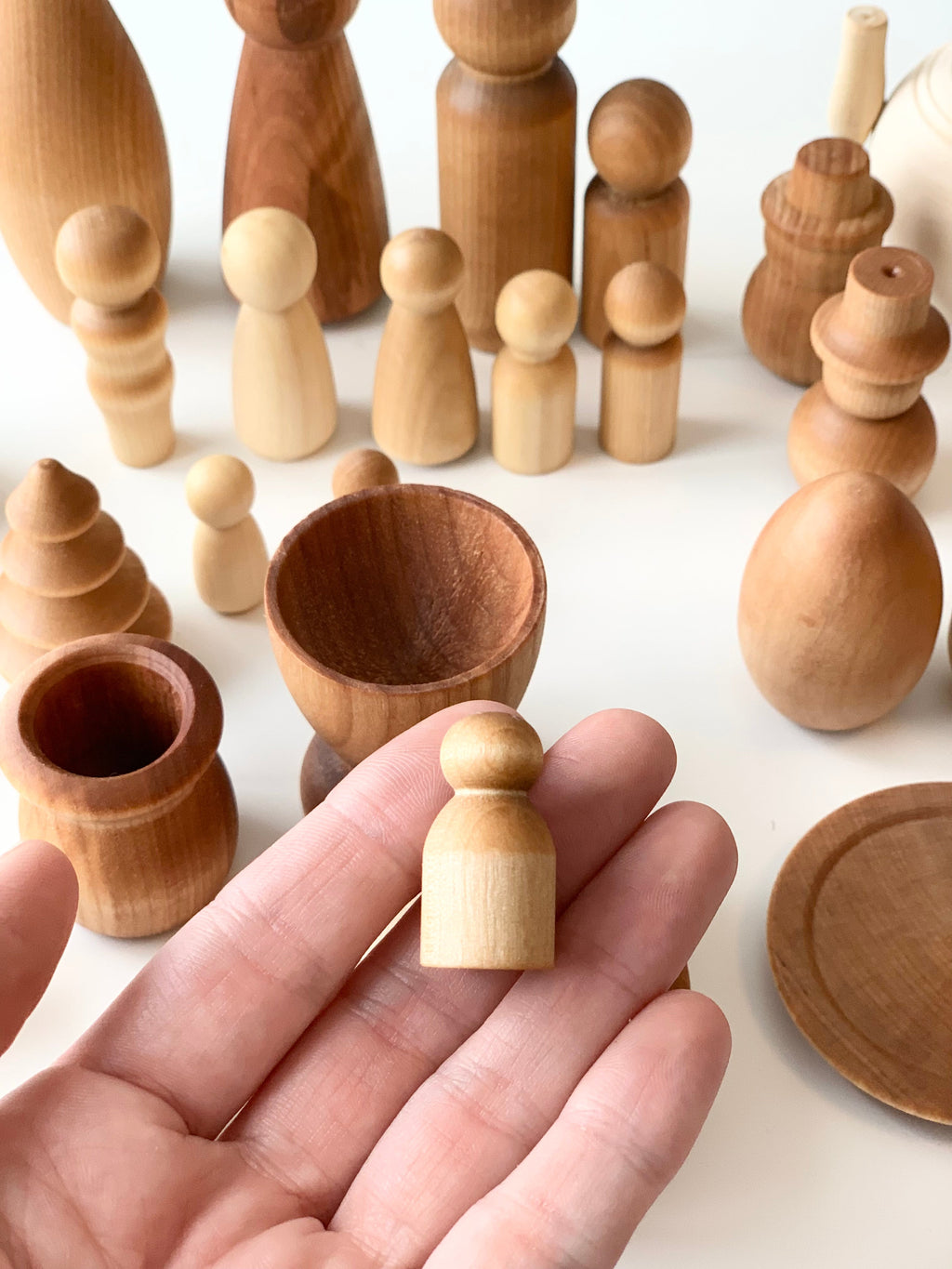 Hand holding small unfinished wooden peg doll among various natural wood shapes and vessels on white surface