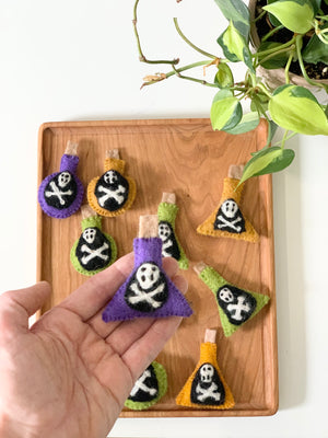 Hand holding a purple felt potion bottle with skull and crossbones on wooden tray with green plant nearby