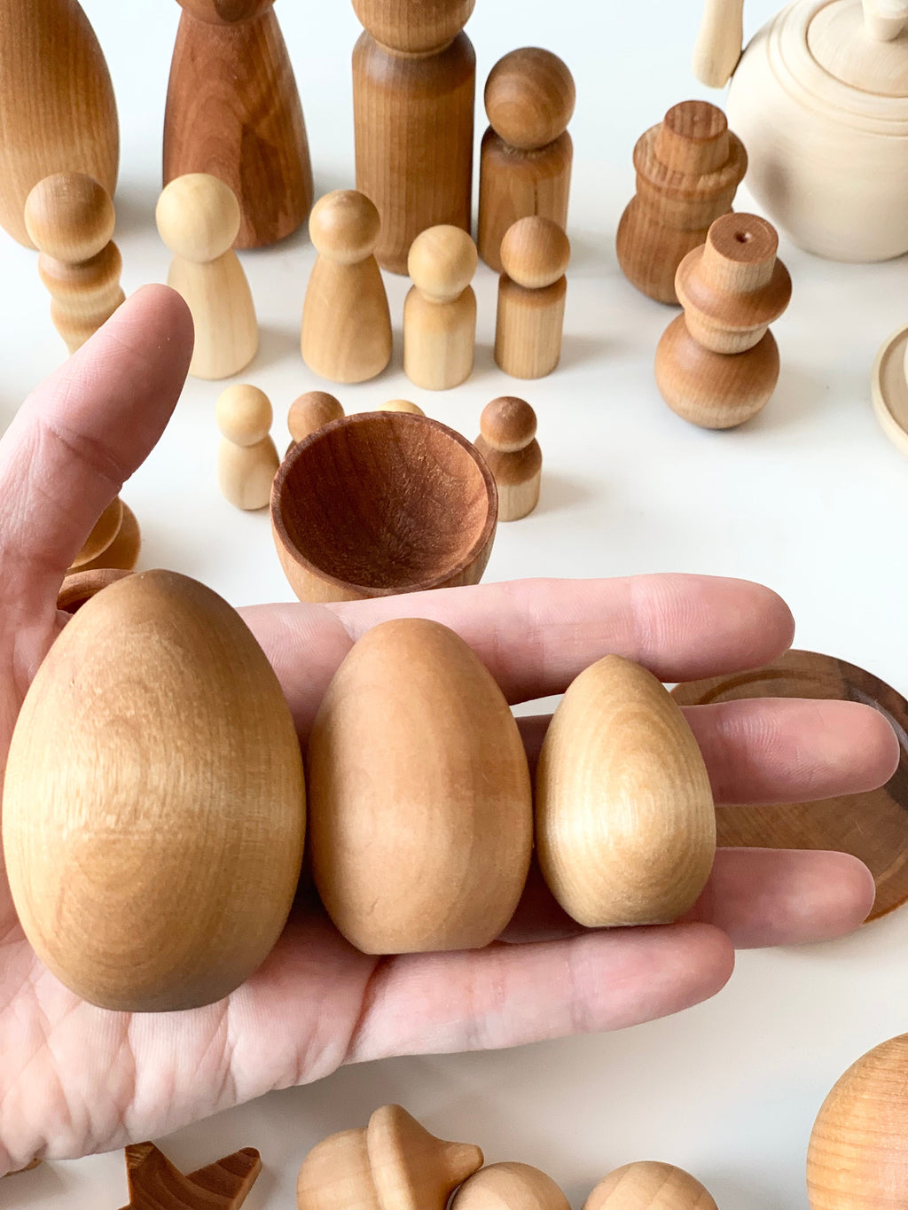 Hand holding three smooth wooden egg-shaped pieces with unfinished wooden figurines and bowls in the background