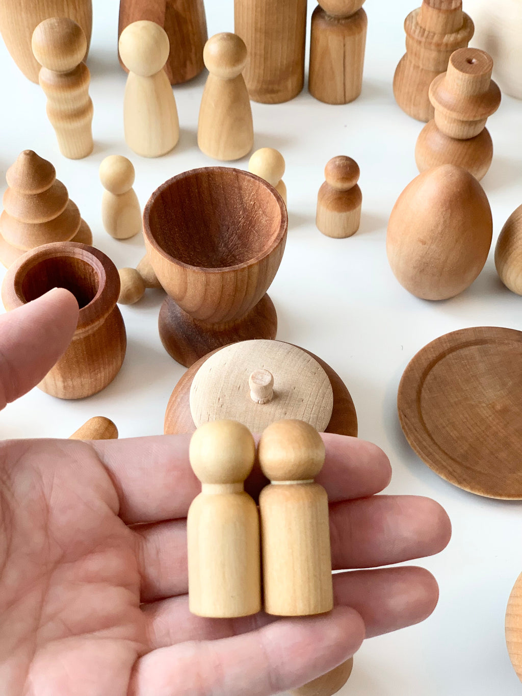 Hand holding two small natural wooden peg figures above various smooth wooden shapes and vessels on white surface