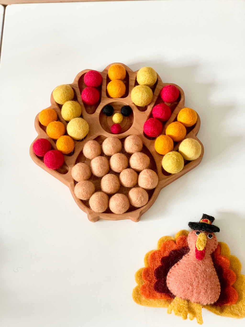Wooden turkey-shaped sensory tray filled with orange, yellow, red, and brown felt balls on white table with felt turkey craft