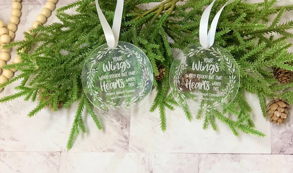 Two round clear acrylic memorial Christmas ornaments with white engraved text and decorative leaves hanging on green pine branches
