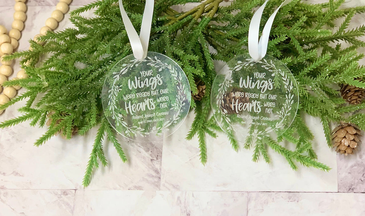 Two round clear acrylic memorial Christmas ornaments with white engraved text and decorative leaves hanging on green pine branches