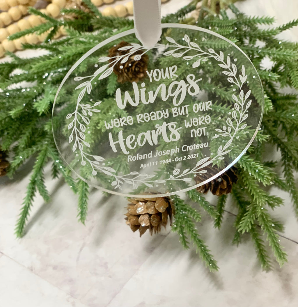 Round clear acrylic memorial Christmas ornament with engraved white floral wreath and sentimental text hanging on evergreen branch