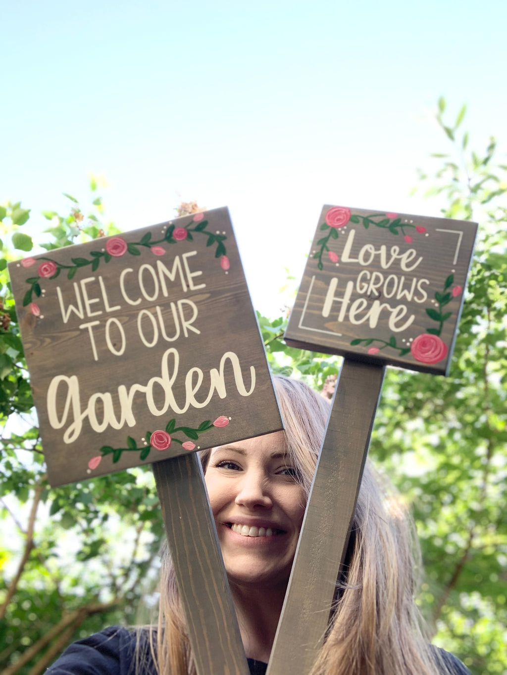 Small square wooden garden signs with white cursive text and pink floral accents held outdoors by a smiling woman