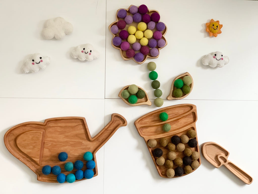 Wooden watering can and flowerpot sensory trays with felt balls arranged as water and flower, plus felt clouds and sun on white surface