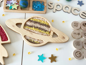 Natural wooden Saturn-shaped sensory tray filled with colored rice on white background with space-themed toys and letters