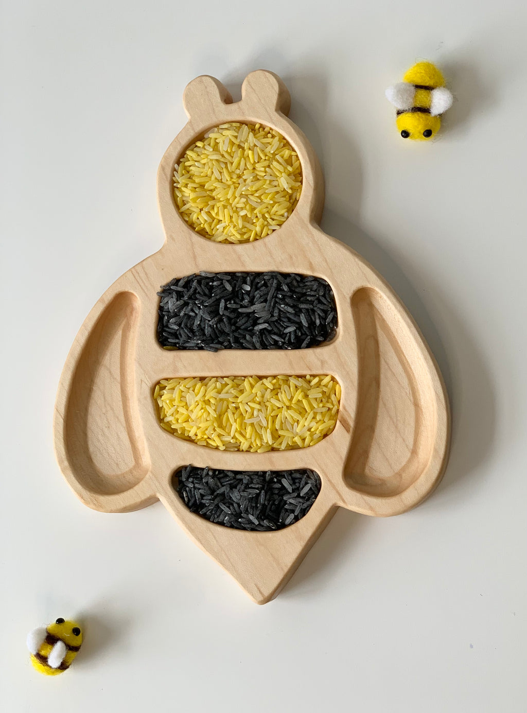 Natural wooden bee-shaped sensory tray filled with yellow and black rice with small bee decorations on white background