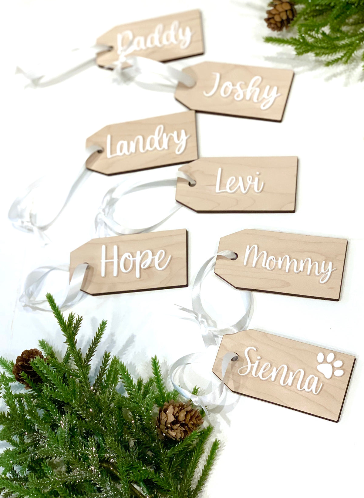 Custom natural wooden Christmas stocking and gift tags with white cursive names and clear ribbon on white background with pine decor