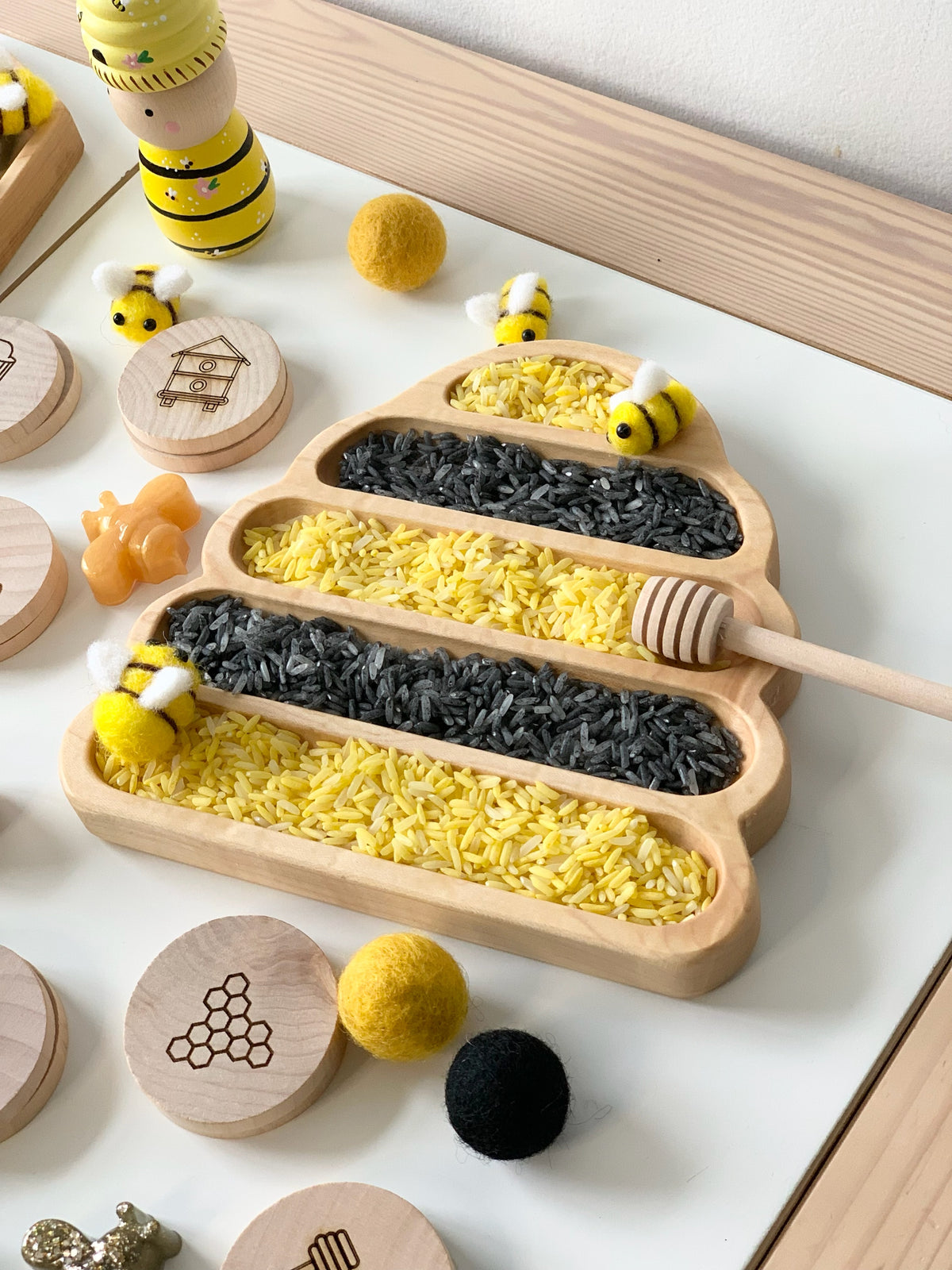 Bee hive shaped wooden sensory tray filled with yellow and black rice and small bee toys on white surface