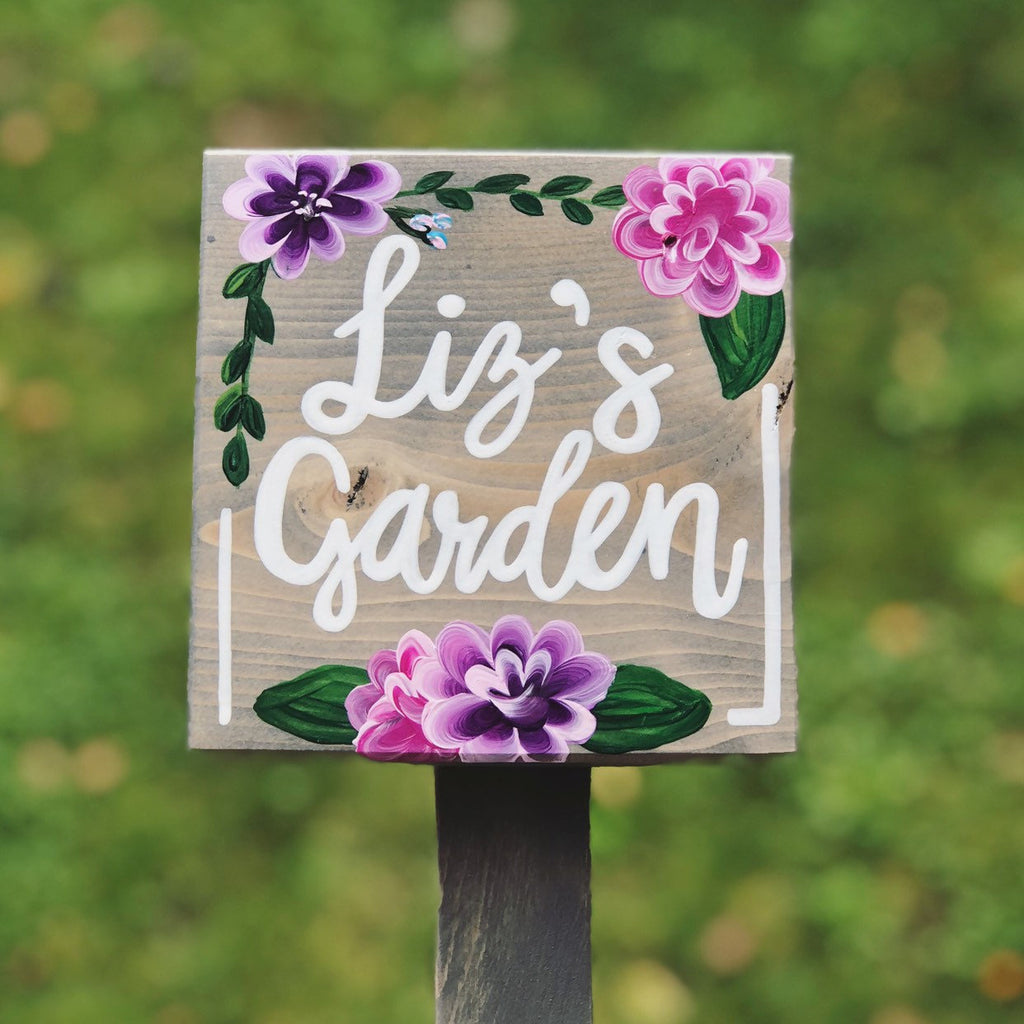 Square wooden garden sign with white cursive letters and hand-painted pink and purple flowers on natural wood background