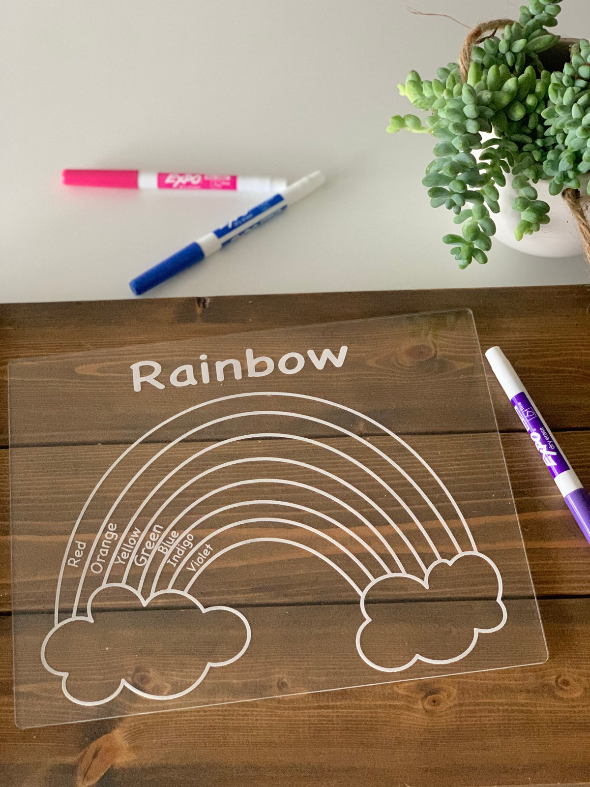 Clear acrylic tracing board with engraved rainbow and color names on wood surface with dry erase markers and plant nearby