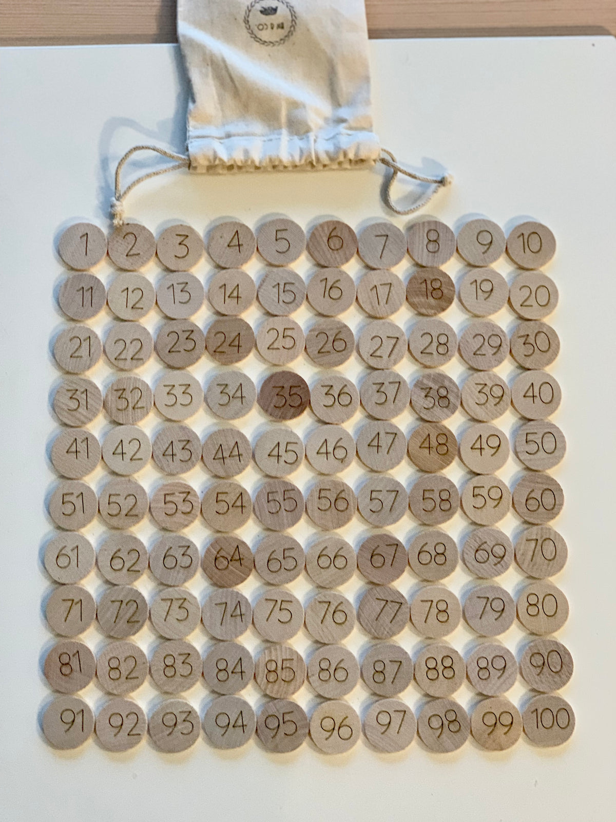 Set of 100 round wooden number tokens from 1 to 100 with drawstring storage bag on white surface