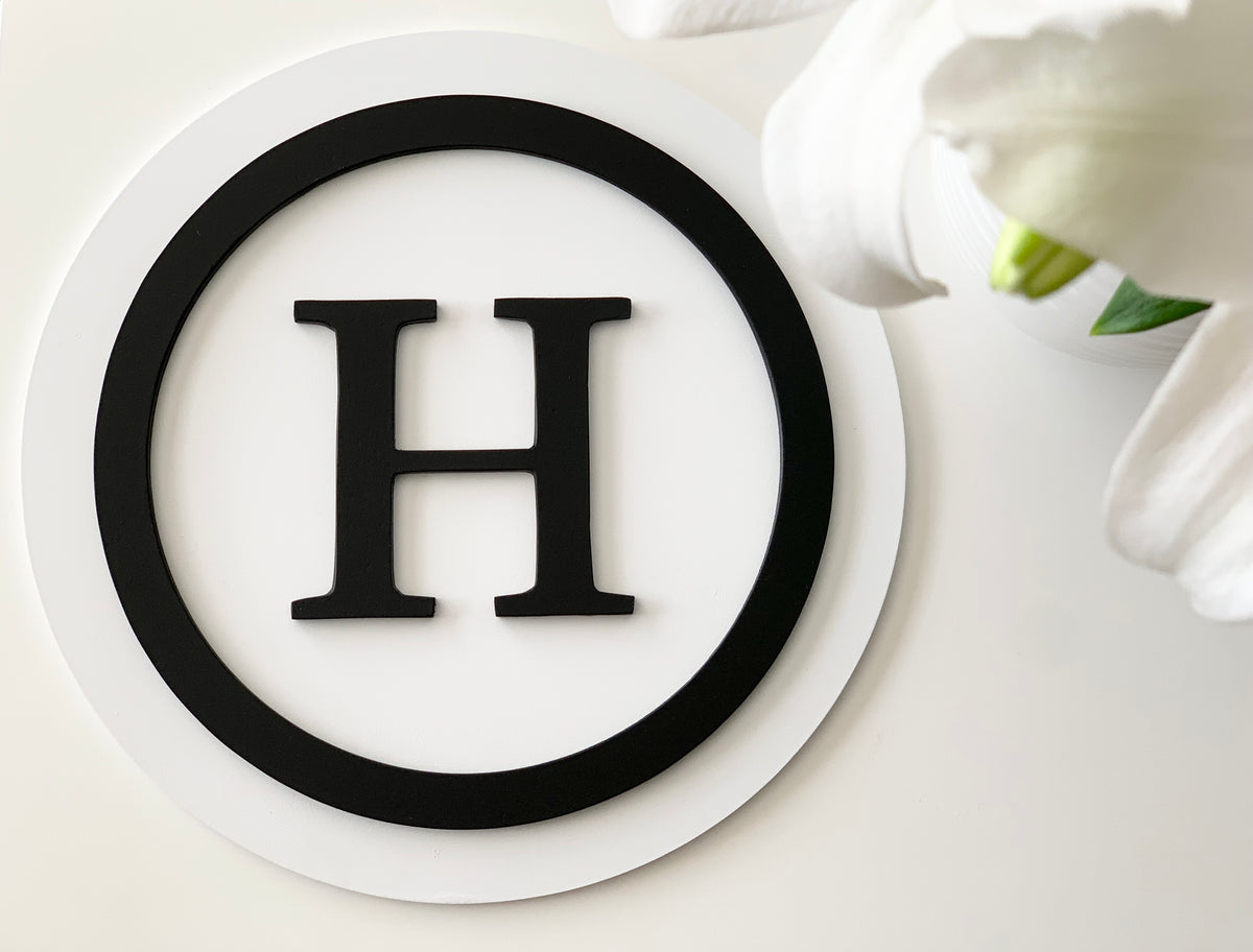 Round 3D monogram sign with black letter H and circle on white two-layer MDF background with white flower accents