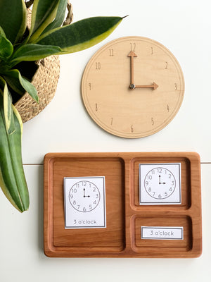 Natural wooden play clock with movable hands and matching time cards on divided wooden tray beside green plant on white surface