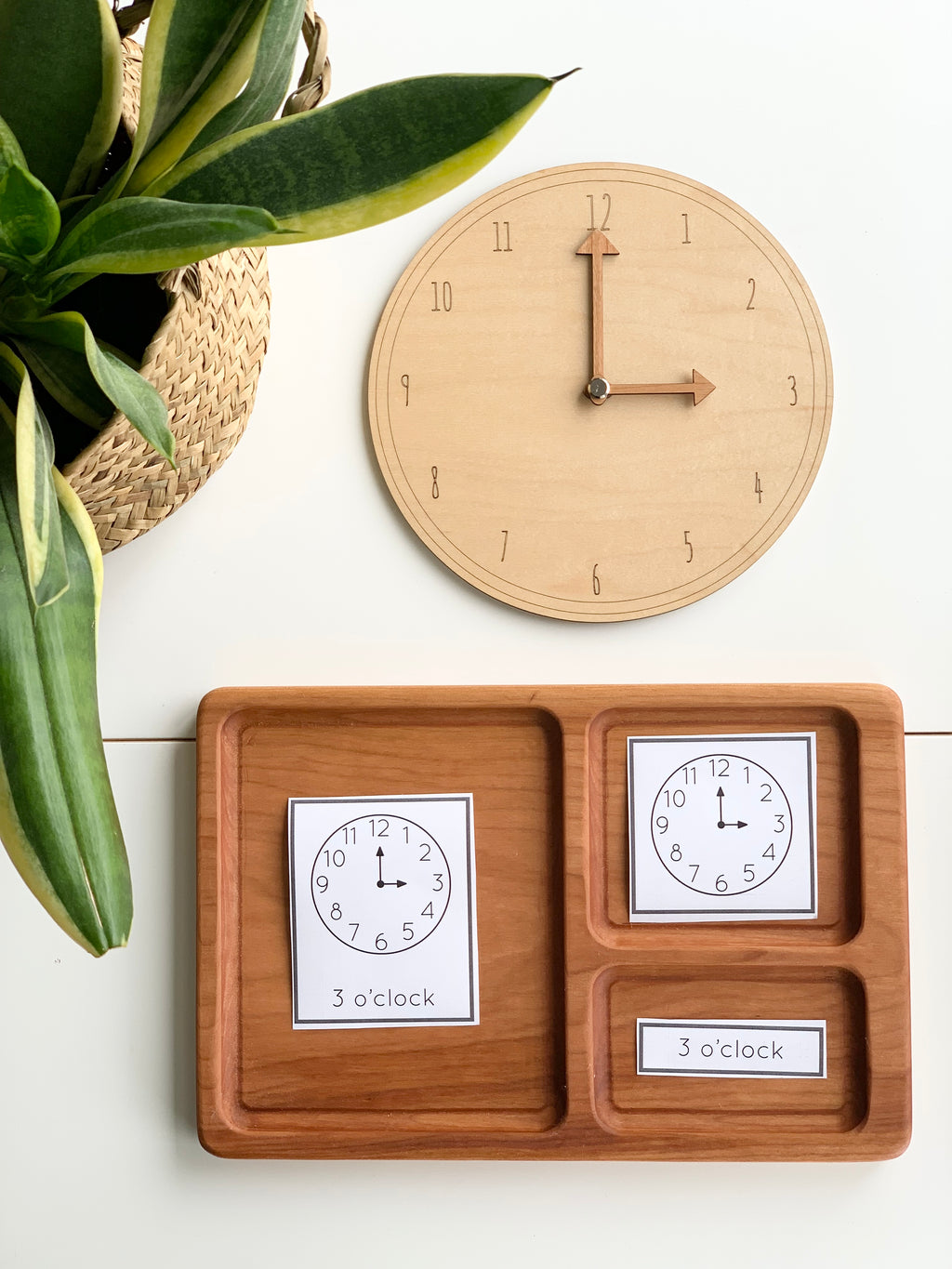 Natural wooden play clock with movable hands and matching time cards on divided wooden tray beside green plant on white surface
