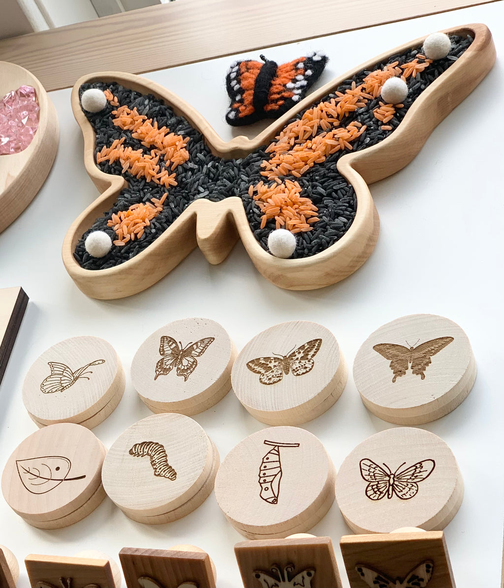 Butterfly-shaped wooden sensory tray with black and orange rice and white pom-poms on light wood surface