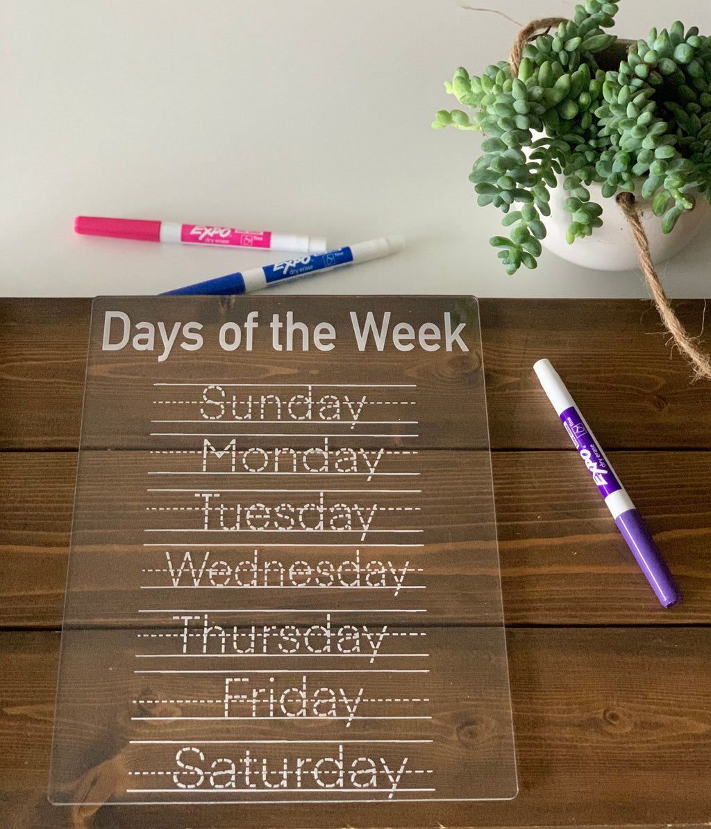 Clear acrylic dry erase board with days of the week traced in dotted letters with dry erase markers on wood table