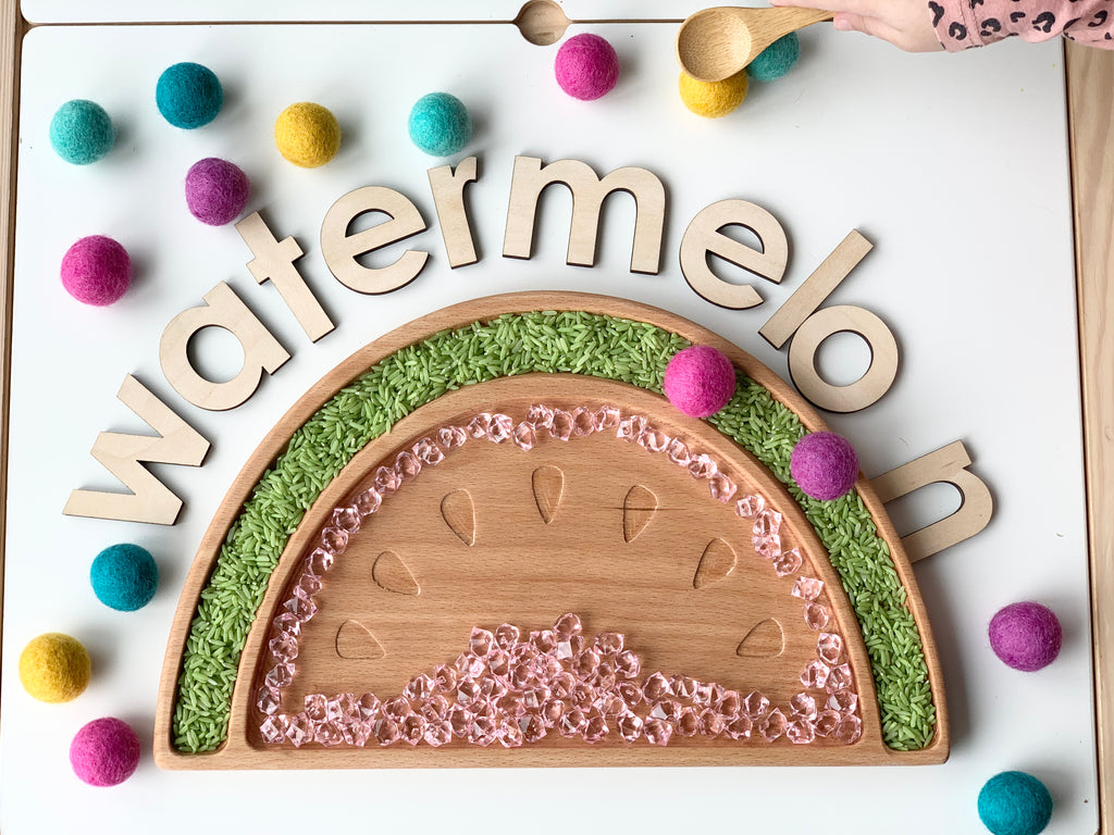 Wooden sensory tray shaped like a watermelon slice with clear beads and colorful felt balls on white surface