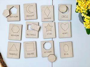 Natural wooden shape puzzle flash cards with engraved shapes and names arranged on white surface with yellow flowers nearby