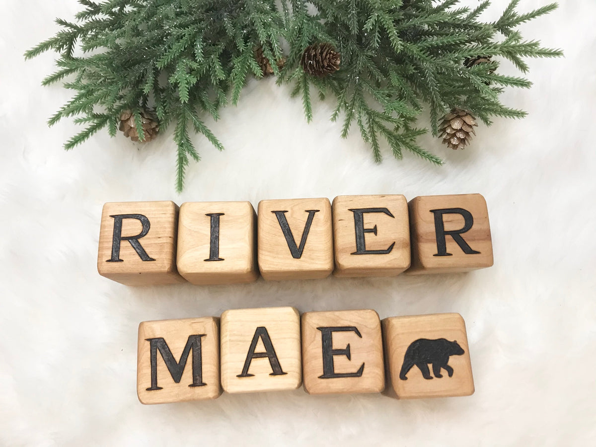 Custom wooden blocks spelling "RIVER" and "MAE" with a bear icon, decorated with pine branches and cones