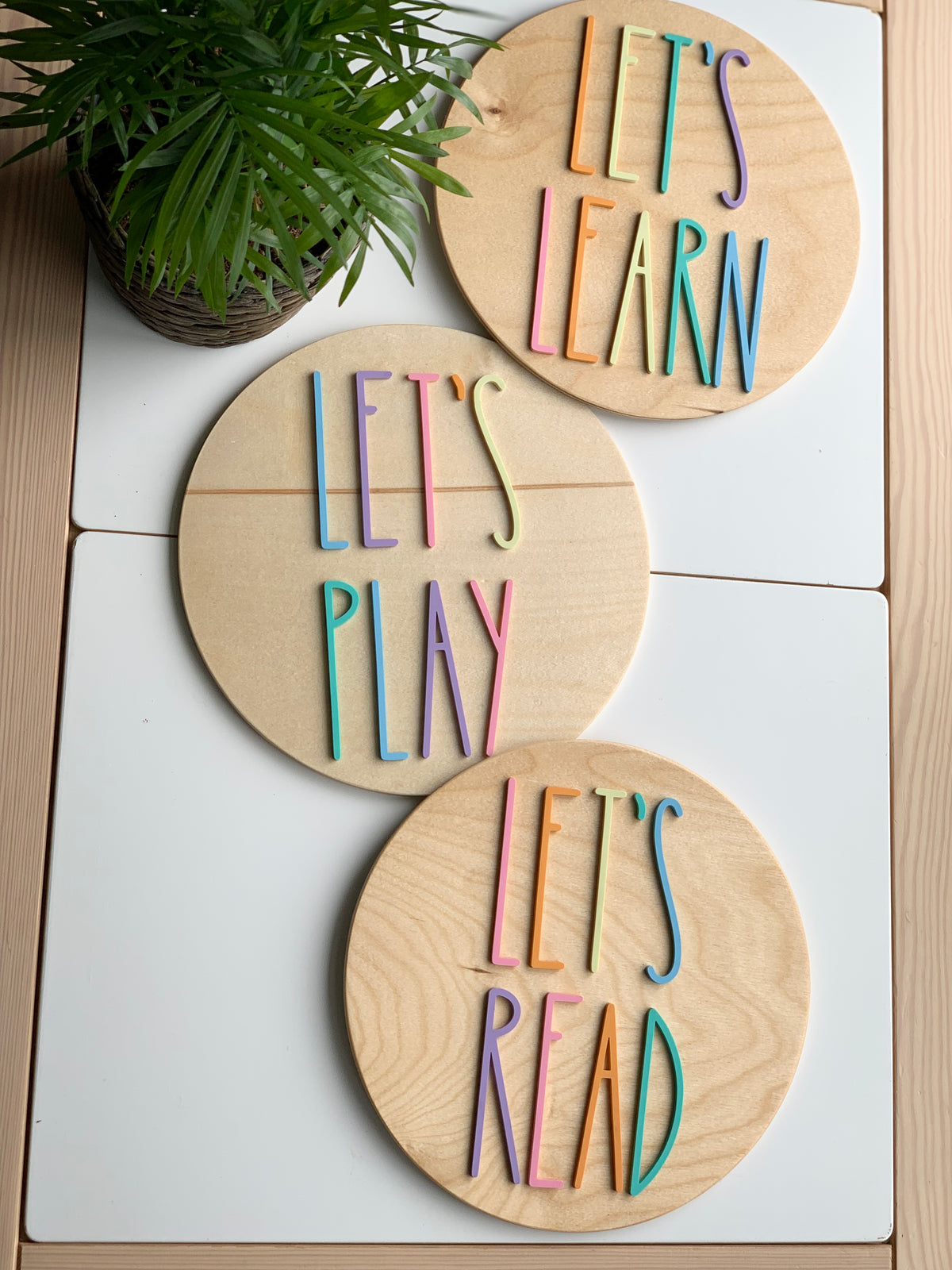 Set of three round wooden signs with colorful letters spelling Let’s Play, Let’s Read, and Let’s Learn on white surface