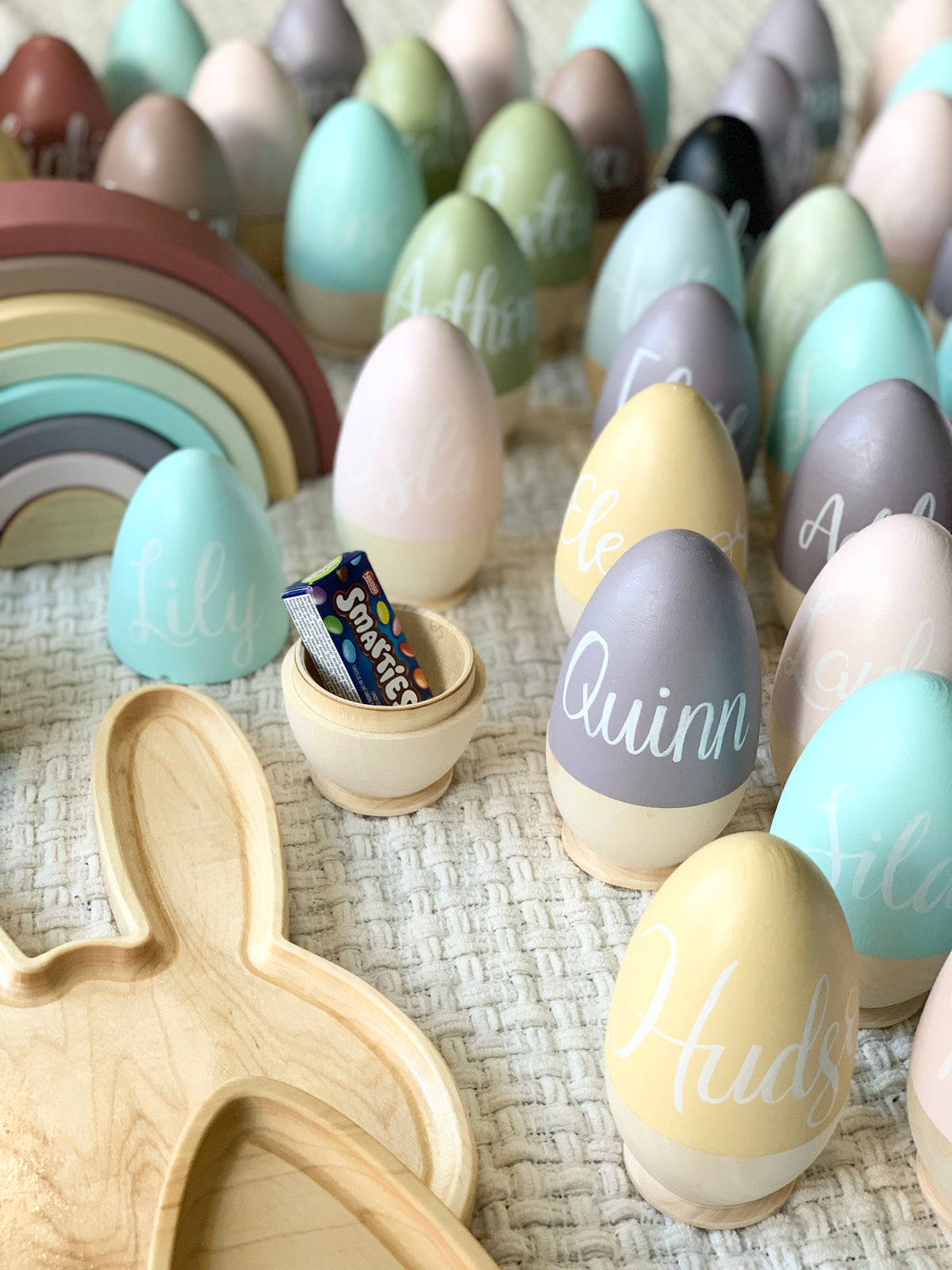 Assorted painted hollow wooden Easter eggs with white cursive names and one open egg showing candy inside on textured fabric