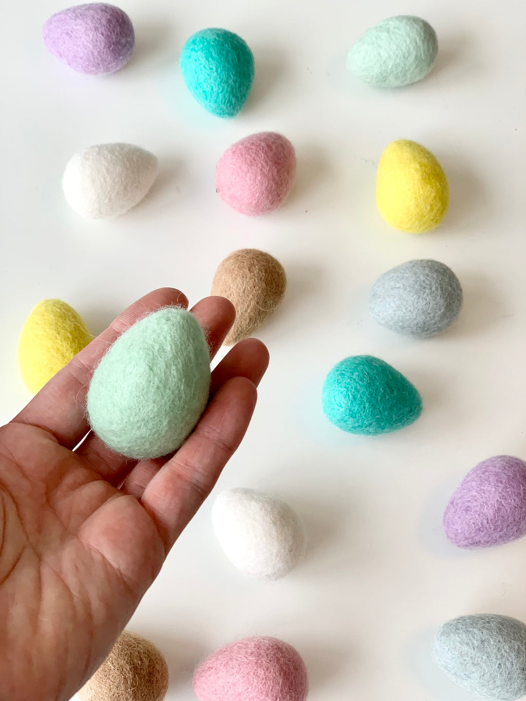 Hand holding a single light green felt Easter egg with scattered pastel-colored felt eggs on white surface